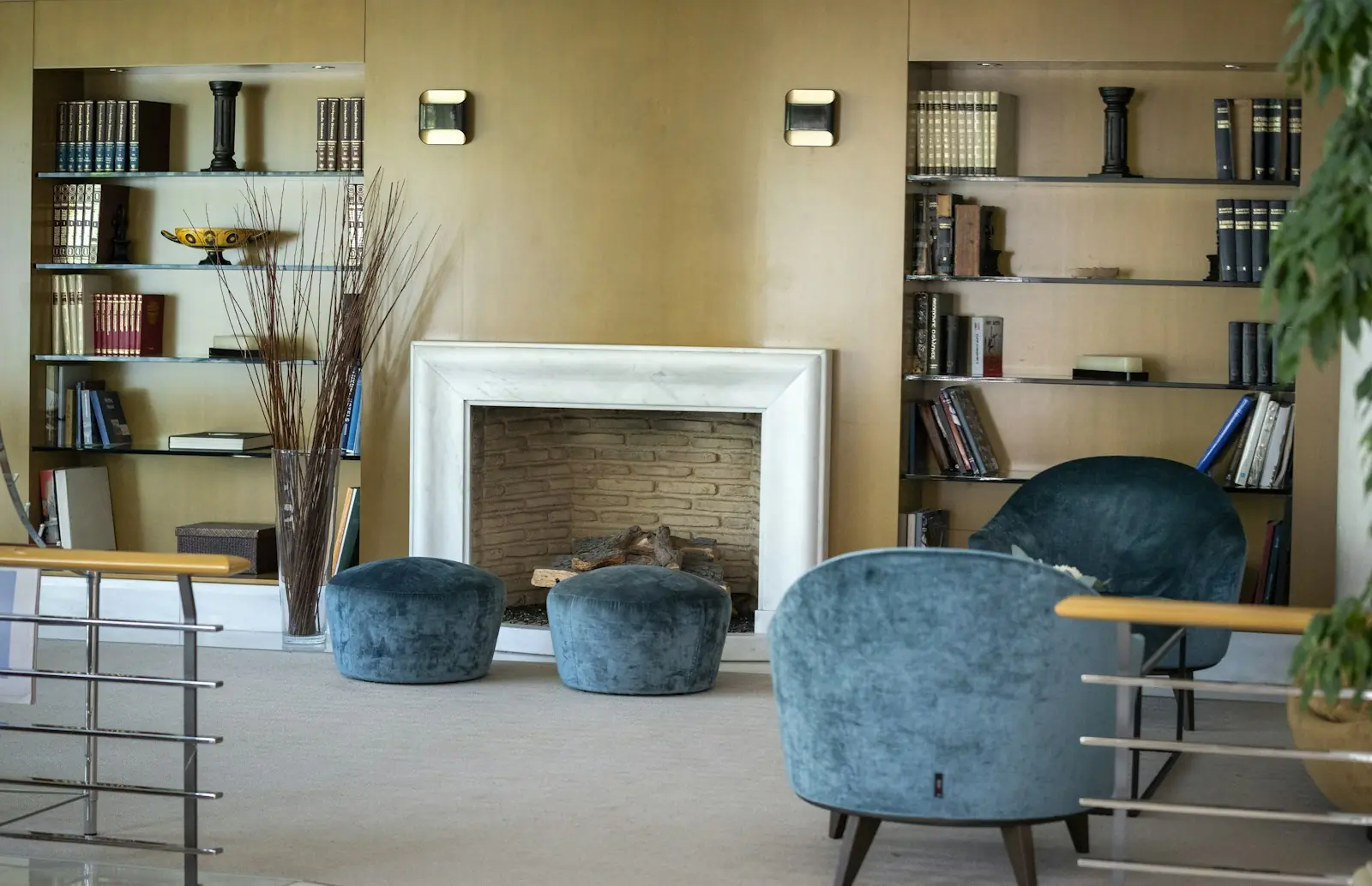 Sala com lareira de mármore branco entre estantes de livros, pufes e poltronas de veludo azul