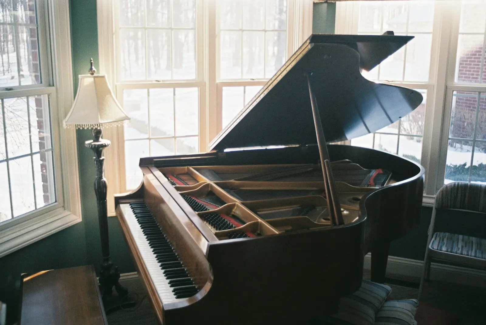 Piano de cauda aberto numa sala pequena, junto às janelas, com abajur ao lado
