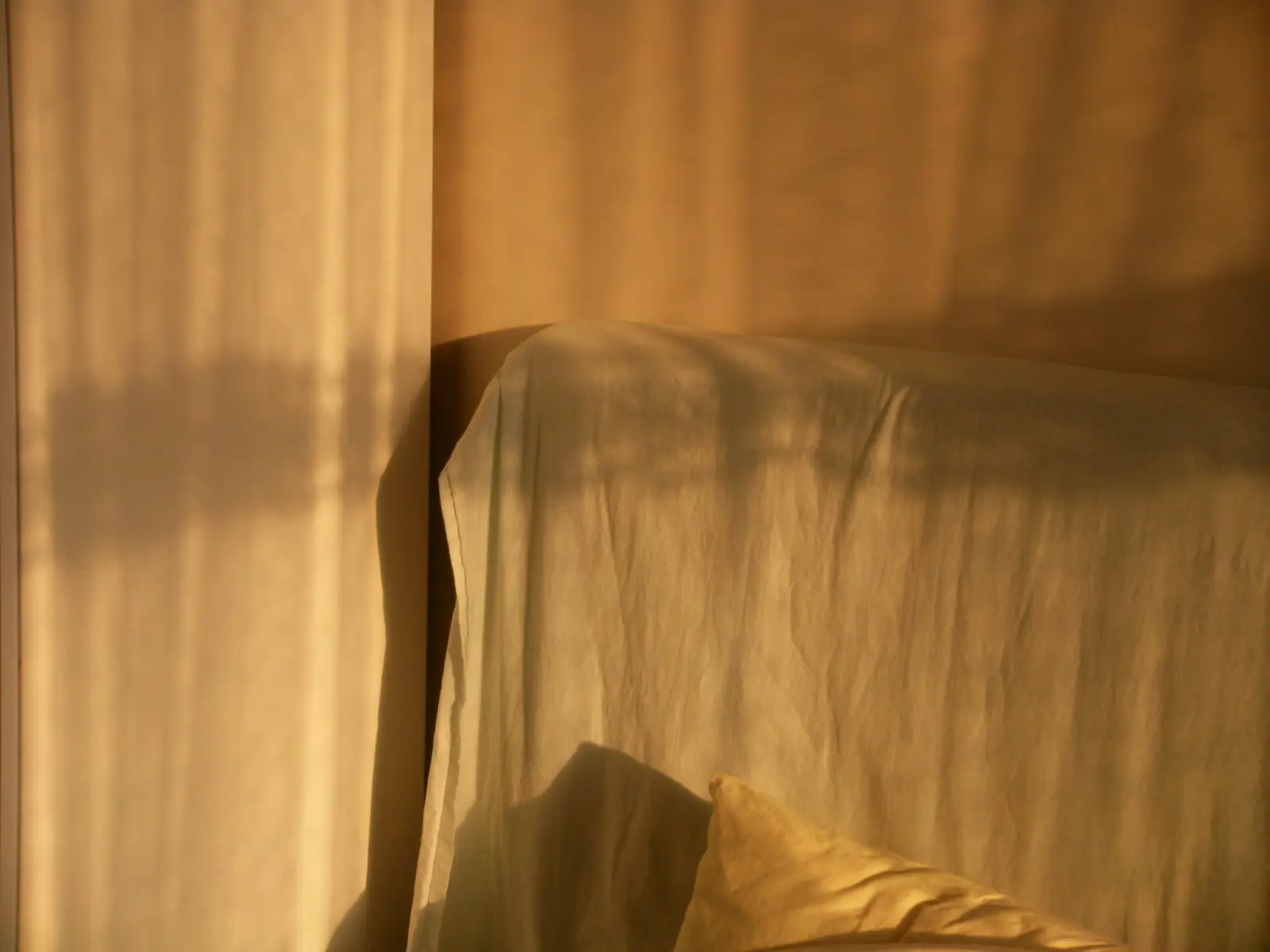 Luz quente atravessando a cortina e desenhando sombras sobre a cabeceira e o lençol de linho