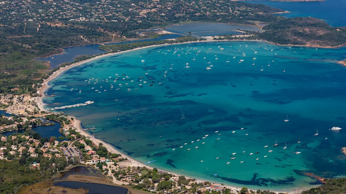 Enseada de águas turquesa com barcos fundeados diante de uma praia em curva, vista do alto